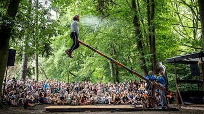 Een acrobaat zit voor een groot publiek bovenaan een schuine balk. Aan de andere zeide van de balk staan muziekanten.
