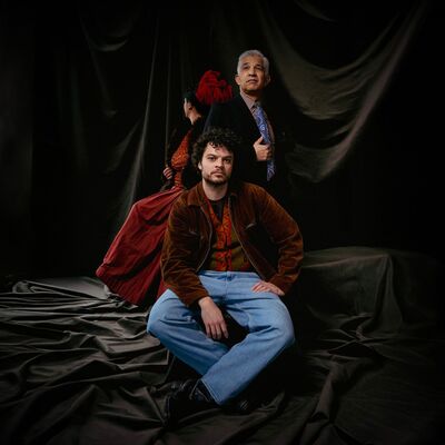 Een groepsportret met drie personen. Op de voorgrond zit een jonge man. Achter hem staat een oudere man. Op de achtergrond staat een vrouw met haar rug naar de camera.
