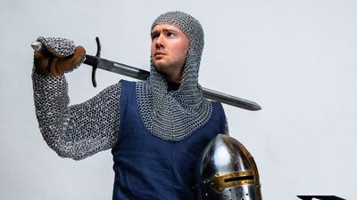 Foto van een ridder, die in de ene hand een helm onder de arm vasthoudt en met de andere hand een zwaard over de eigen schouder legt.