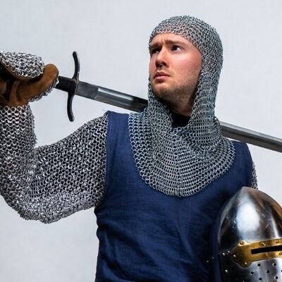 Foto van een ridder, die in de ene hand een helm onder de arm vasthoudt en met de andere hand een zwaard over de eigen schouder legt.